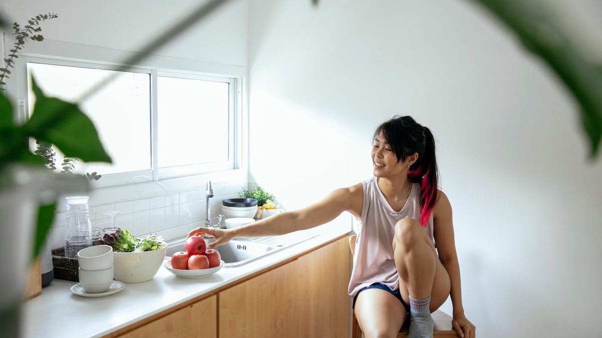 Positive ethnic young lady in pajama sitting near counter with utensil and apples in plate near window in kitchen in daytime